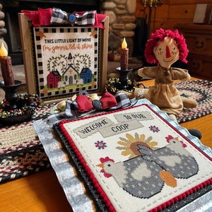 May include: A framed cross-stitch with the text "THIS LITTLE LIGHT OF MINE I'm gonna let it shine" and a depiction of a church and houses. A second framed cross-stitch with the text "WELCOME TO OUR COOP" and a depiction of two chickens. A rag doll with red hair and a brown dress. A wooden table with a woven rug.