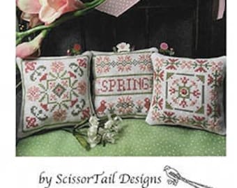 Op de afbeelding: Drie kruissteek kussenontwerpen met een lentethema. De kussens zijn wit met rode, groene en roze accenten. Het middelste kussen heeft het woord "SPRING" in rood geborduurd. De tekst "Spring on the Square" staat bovenaan de afbeelding.