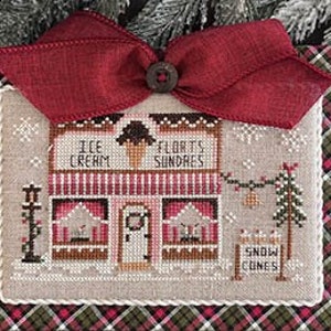 Op de afbeelding: Kruissteekpatroon van een kleine, rood-witte winkel met een bord waarop "Ice Cream & Florets Sundaes" staat. De winkel heeft een rode deur en twee ramen met een kleine kerstboom en een bord waarop "Snow Cones" staat. Het patroon is omlijst met een rood-groene ruit.