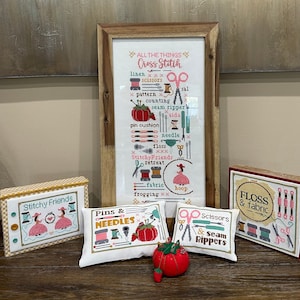 May include: A framed cross stitch design titled "All the Things Cross Stitch" with a list of cross stitch supplies including linen, scissors, pattern, counting, seam ripper, aida, pin cushion, needle, floss, retreat, fabric, frogging, and hoop. There are also four smaller cross stitch projects: "Stitchy Friends", "Pins & Needles", "Scissors & Seam Rippers", and "Floss & Fabric".