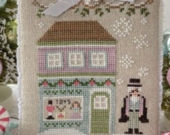 May include: A cross-stitch pattern of a toy shop with a pink facade, green trim, and a sign that reads "TOYS". A snow-covered roof with a white ribbon and a snowman in a top hat stand outside the shop.