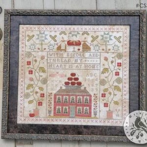 May include: Framed cross-stitch artwork featuring a house with a stack of apples on top, surrounded by floral patterns. The text "WITH NEEDLE & THREAD MY HEART IS AT HOME" is displayed. The artwork is in a dark wooden frame.
