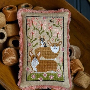 May include: A cross-stitch sampler featuring three Corgi dogs in a garden setting with pink flowers. The sampler is on a pillow with a pink velvet trim. The title of the sampler is "Corgi Caboodle".