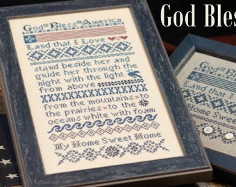 May include: Two framed cross-stitch embroidery pieces with the text "God Bless America" and "Land that I Love" in blue and red thread. The frames are dark wood and the background fabric is a natural linen colour.