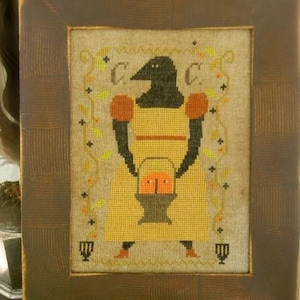 May include: A framed cross-stitch embroidery depicting a black crow wearing a yellow dress and holding a basket of orange pumpkins. The embroidery is surrounded by a brown wooden frame with a textured surface.