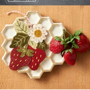 May include: A decorative ornament featuring felt strawberries, leaves, and a daisy on a honeycomb-shaped base. The ornament is approximately 4 inches by 4.5 inches. The text "Sweet Berries Ornament" is visible.