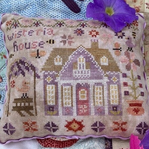 May include: A small, hand-stitched pillow with a purple and brown house design and the text "Wisteria House". The pillow is on a patchwork quilt with purple and pink flowers.