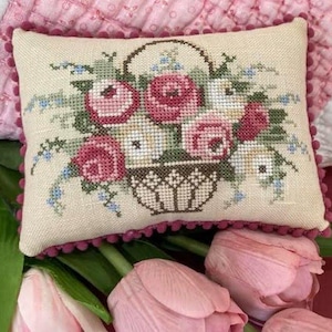 May include: A small, cream-colored pillow with a pink trim features a cross-stitch design of a basket overflowing with pink and white roses. The pillow is sitting on a pink and white quilt with a bouquet of pink tulips in the foreground.