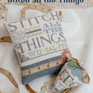May include: A cross-stitch project with the text "Stitch All the Things" on a blue and white pillow. The pillow is surrounded by a wooden embroidery hoop, buttons, and a measuring tape.