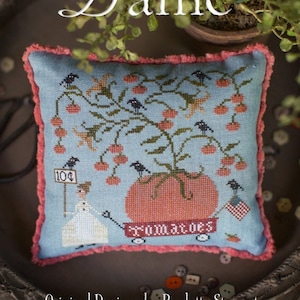 May include: A blue and red cross-stitch sampler pillow with a tomato plant, birds, and a woman pushing a red wagon with the word "tomatoes" on it. The pillow is on a wooden tray with a green plant in a brown pot. The text "Plum Street Samplers" and "Tomato Dame" are visible above the pillow.