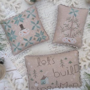 May include: Three beige cross-stitch pillows with winter themes. One pillow features a snowman with a top hat, another a tree, and the third reads "let's build snowman." The image includes pine needles, ornaments, and the text "Snowmen in the Forecast."