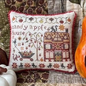 May include: A white pillow with red trim and a cross-stitch design depicting a house with the text "candy apple house". The pillow is sitting on a patchwork quilt with a carved pumpkin in the background.