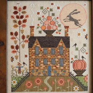 May include: Framed cross-stitch sampler titled "Autumn Moon" by Plum Street Samplers. The artwork depicts a brick house with a pumpkin, a running hare, and a full moon. The frame is dark wood.