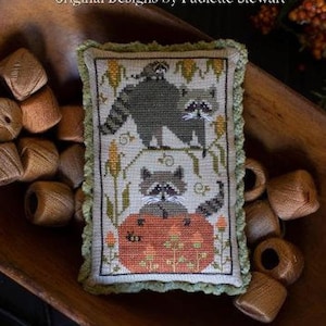 May include: A cross-stitch sampler pillow featuring a design of three raccoons with a pumpkin. The pillow is in a wooden bowl with spools of thread. The text on the pillow reads "Raccoon Rabble".
