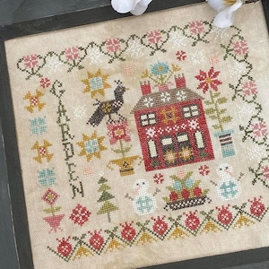 May include: A framed cross-stitch sampler with a red house, a bird, and the word "Garden" in a floral design. The sampler is on a colorful patchwork quilt with a white bird figurine and a snowman.