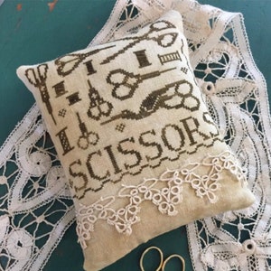 May include: A beige needlepoint pillow with a brown embroidered design of scissors and the text "I Love Scissors". The pillow is trimmed with white lace and is sitting on a teal surface with a white lace doily.