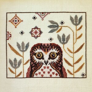 May include: Framed cross-stitch artwork featuring a detailed owl with large yellow eyes, brown and white feathers, and a red and white chest. The owl is surrounded by stylized flowers and geometric patterns in a cream-colored background. The frame is dark brown.