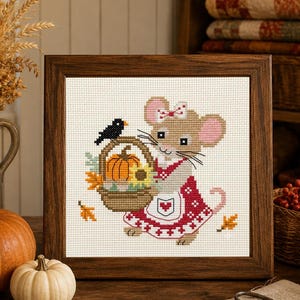 May include: A framed cross-stitch embroidery featuring a tan mouse wearing a red and white patterned dress and apron, holding a woven basket filled with an orange pumpkin and a yellow sunflower, with a small black bird perched on the handle.