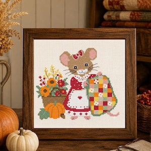 May include: A framed cross-stitch embroidery featuring a brown mouse wearing a red dress and white apron, holding a colorful patchwork quilt. Beside the mouse is a basket of autumn flowers and a bright orange pumpkin on a cream background.