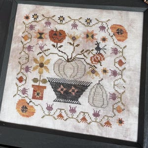 May include: A framed cross-stitch featuring pumpkins, flowers, and a witch's hat in autumnal colors. The black frame sits on a quilted fabric with orange, brown, and beige patterns. Two knitted pumpkins and scissors are also present.