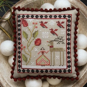 May include: A cross-stitch sampler with a winter scene featuring a woman, a deer, birds, and a house. The sampler is framed in a red and black border and is displayed on a white pillow. The pillow is sitting in a bowl with white pom-poms. The text "Winter Blessings" is displayed below the image.