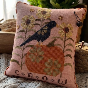 May include: A pink pillow with a cross-stitch design of a black bird perched on a pumpkin with the word "CROW" stitched below. The pillow is on a white knitted blanket with a bouquet of orange flowers in the background. The text "In The Pumpkin Patch" is at the top of the image and "Designed by Annie Belcher" is at the bottom.