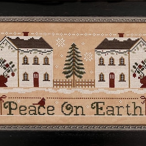 May include: Cross-stitch pattern featuring two white houses with red doors, a green evergreen tree, and red berry bushes. The text "Peace on Earth" is stitched below the houses.