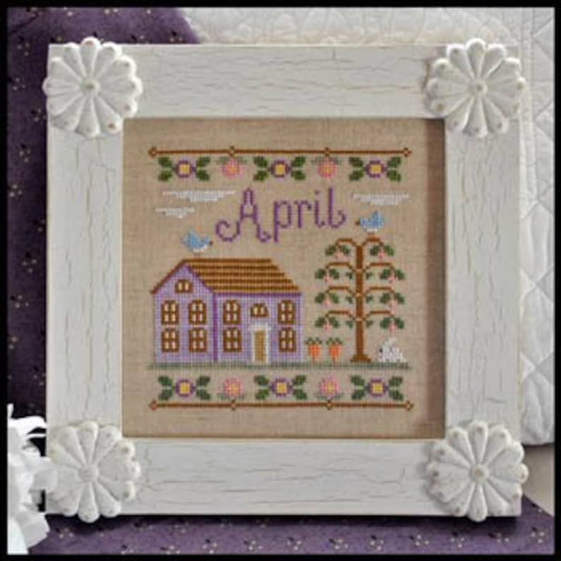 K&ouml;nnte beinhalten: Kreuzstich-Stickerei-Design mit einem lila Haus mit braunem Dach, einem Baum mit gr&uuml;nen Bl&auml;ttern und einem wei&szlig;en Kaninchen. Das Design ist in einem wei&szlig;en Rahmen mit floralen Akzenten gerahmt und das Wort "April" ist in der Mitte gestickt.