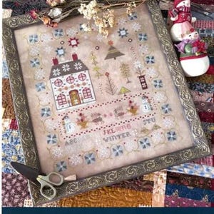 May include: An embroidered sampler titled "Enchanted Winter Sampler" with a house, trees, and snowflakes. The design includes the text "ABC WINTER STU" and is framed in a decorative gold frame. The image also includes scissors and a snowman figurine.
