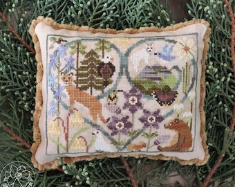 May include: A decorative, rectangular cross-stitch pillow featuring a nature scene with a deer, bear, and other animals. The design is framed by a heart shape with blue and purple flowers, and a tan fringe border.