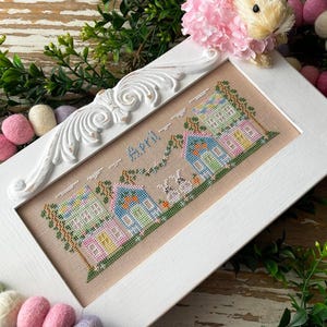 May include: A white framed cross-stitch embroidery with a pastel colored scene of houses and bunnies. The embroidery is titled "April" and is surrounded by a garland of pastel colored felt balls.
