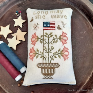 May include: A white fabric pillow with a cross-stitch design featuring a potted plant with red flowers and an American flag. The text "Long may she wave" is stitched above the plant.