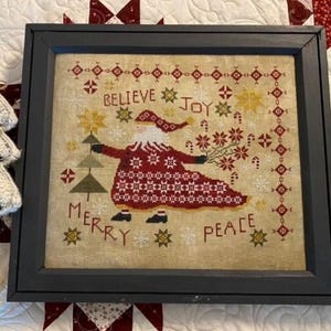 May include: A framed cross-stitch sampler with a red and white Santa Claus figure holding a sprig of holly. The sampler features the words "Believe", "Joy", "Merry", and "Peace" in a vintage style font.
