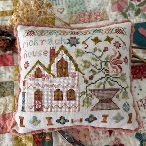 May include: A small, square, white pillow with a cross-stitched design featuring a house and the words "rick rack house." The design includes floral and geometric elements in shades of pink, green, and brown. The pillow has a pink, ruffled edge.