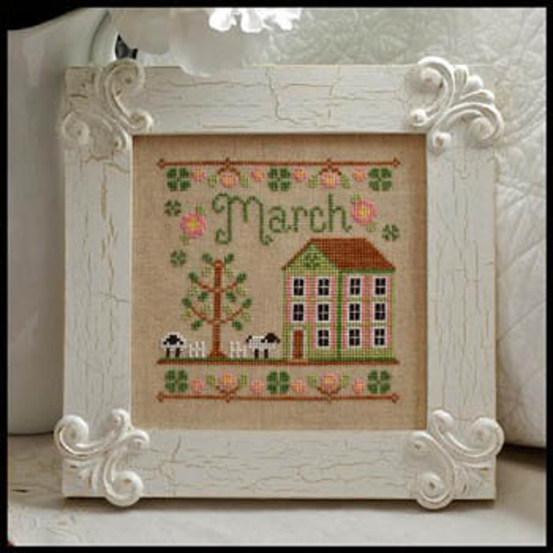 May include: Cross stitch pattern featuring a green house with a white picket fence, a tree, and sheep. The text "March" is stitched in cursive above the house. The background is a light beige fabric.
