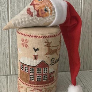 May include: A Christmas-themed decorative item featuring a Santa Claus hat and a cylindrical base. The base is adorned with cross-stitch images of a house, reindeer, and snowflakes. The Santa hat is red with a white trim and a fluffy white pom-pom.