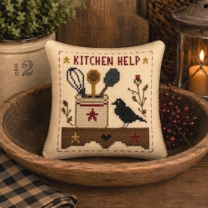 May include: A square fabric pincushion featuring a cross-stitch design with the text KITCHEN HELP. The embroidery depicts a white crock filled with a whisk, wooden spoon, and spatula, alongside a black bird, floral sprigs, and a brown shelf.