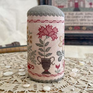 May include: A cylindrical pincushion featuring cross-stitch embroidery on light pink fabric. The design displays a central dark brown urn holding a large pink flower, surrounded by smaller floral motifs in muted tones. It has a grey scalloped trim.