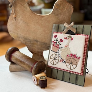 May include: A wooden rooster figurine and a framed cross-stitch picture of a goose riding a bicycle with flower baskets. The frame is green with a red and white checkered border. Also, a large wooden spool and two spools of thread.