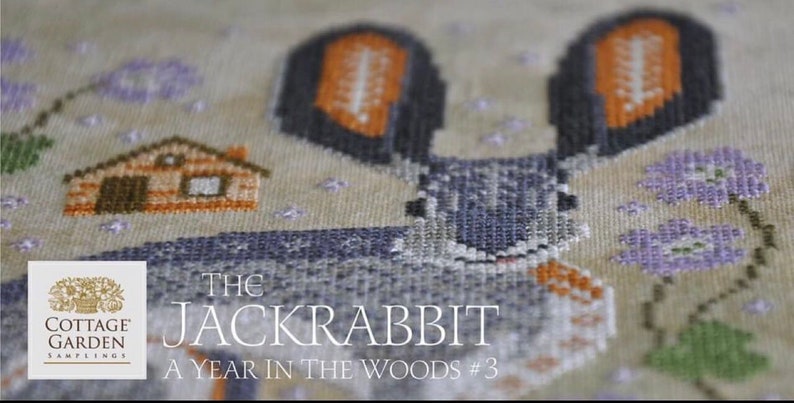 May include: Cross stitch pattern featuring a gray rabbit with orange ears, a small wooden house, and purple flowers. The text reads "The Jackrabbit A Year in the Woods #3" and "Cottage Garden Samplings".