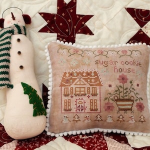 May include: A white and brown embroidered pillow with a gingerbread house design and the text "sugar cookie house". The pillow is surrounded by white pom-poms and is sitting on a red and white quilt.
