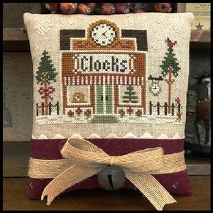 May include: A beige pillow with a brown and tan embroidered design of a clock shop with the word "Clocks" above the door. The pillow has a burgundy fabric backing with a brown ribbon and a bell.