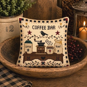 May include: A square decorative pillow featuring a cross-stitch design on a cream background. The text COFFEE BAR is centered above a brown table holding a coffee grinder, a mug, and two canisters labeled COFFEE and BEANS. A black bird sits above.