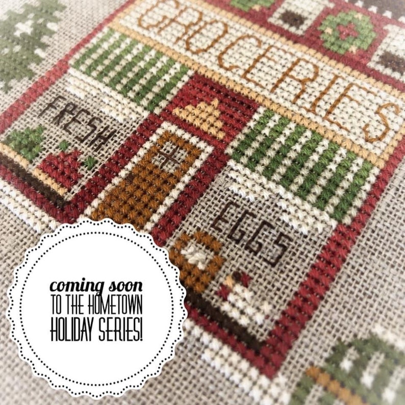May include: Cross-stitch pattern of a red and green grocery store with the word "GROCERIES" above the door and "EGGS" below the door. The text "FRESH" is visible on the left side of the image. The image is labeled "coming soon to the HOMETOWN HOLIDAY SERIES!"