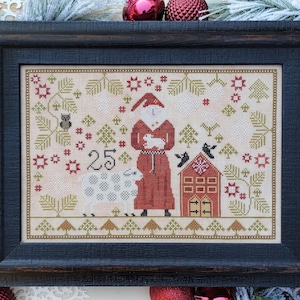 May include: A cross-stitch pattern featuring a red-robed Santa Claus holding a white lamb, a white sheep, a small red house with a white door, and the number 25. The pattern is surrounded by green trees, red stars, and brown branches.