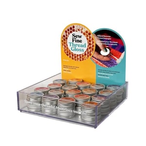 May include: A display case filled with small, silver tins of Sew Fine Thread Gloss. The display features a yellow and blue sign with the product name and a honeycomb design. The text "Tame your threads!" is printed on the case.