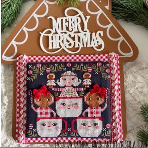 May include: A cross-stitch embroidery on a dark fabric featuring a central figure and two gingerbread characters in mugs, framed by a red and white gingham border on a brown gingerbread house-shaped wooden plaque with the text Merry Christmas.