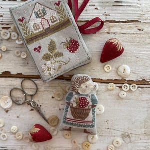 May include: A collection of handmade cross-stitch items. Includes a stitched book cover with a house and strawberry design, a hedgehog holding a basket of strawberries, two strawberry-shaped pin cushions, and various buttons. The text "Animal Cracker Series + Pip+" and "Design by Stacy Nash" are visible.