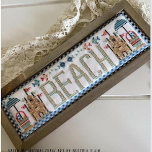 May include: A rectangular cross-stitch sampler in a wooden frame featuring the word BEACH in light green letters. The design includes two sandcastles with blue umbrellas, red flags, and small colorful fish accents on a white fabric with blue gingham trim.