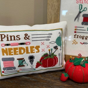 May include: A white pillow with a cross-stitch design that says "Pins & Needles". The design includes a variety of sewing supplies, including needles, pins, and a spool of thread. The pillow is sitting on a wooden table.
